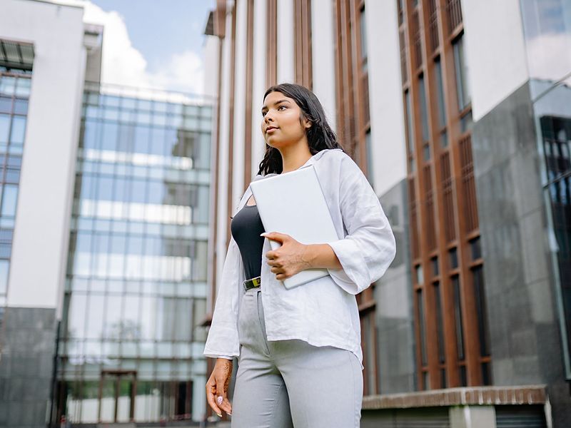 A young woman holding a laptop outside a glass office building.